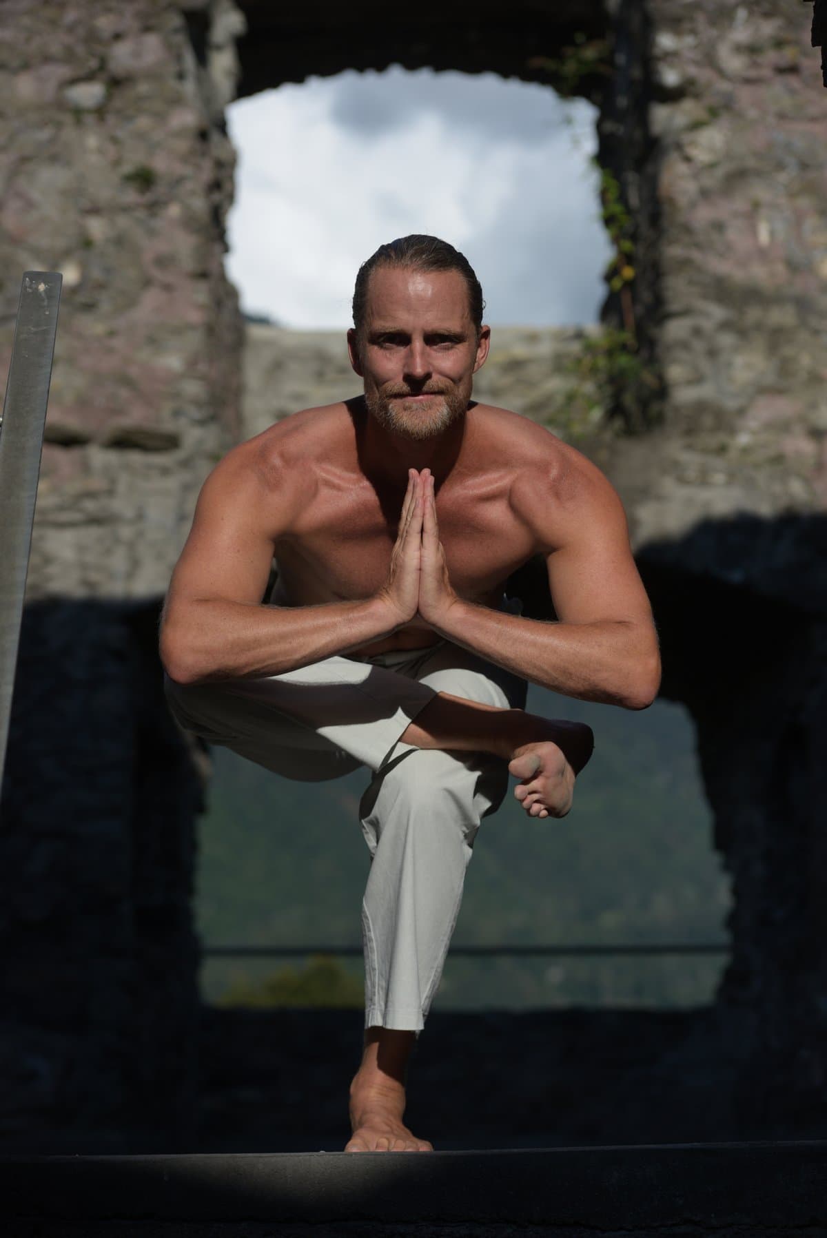 Jason Milne in a standing tree pose with prayer hands, framed by a stone ruin